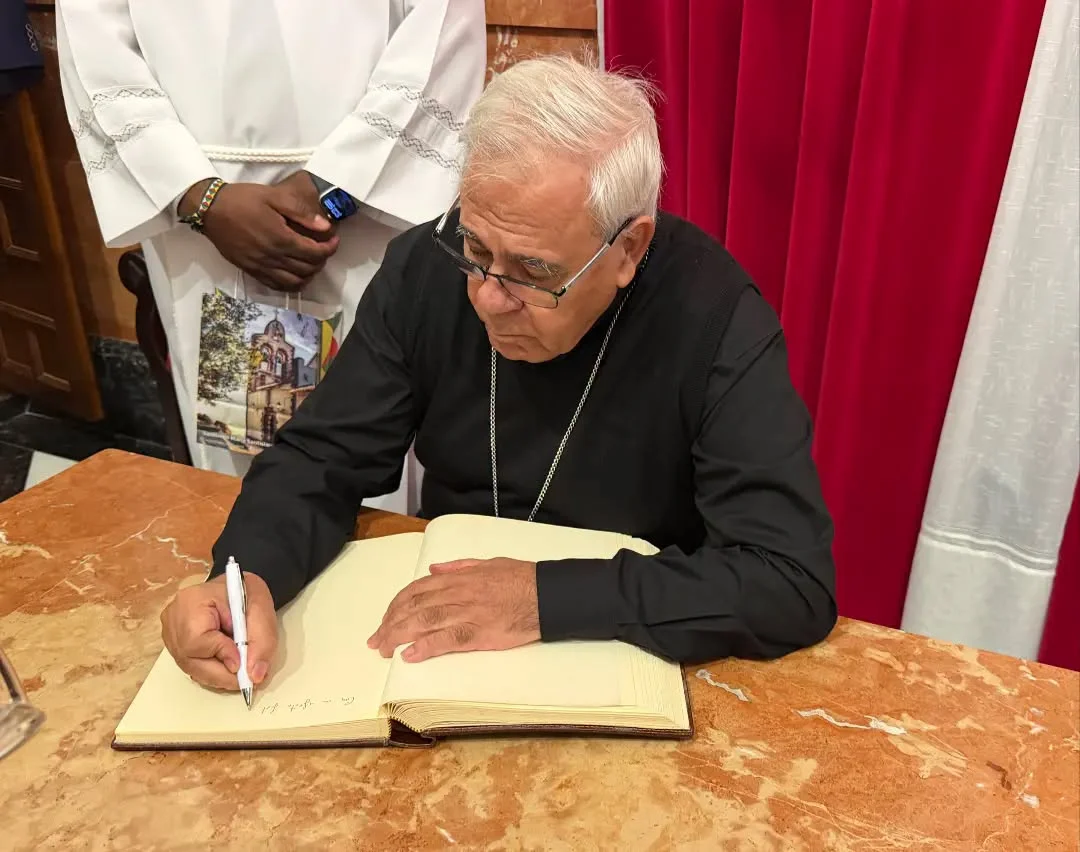 Un hombre mayor con gafas y camisa negra escribe con un bolígrafo en un libro abierto sobre una mesa de mármol, junto a una persona con túnica blanca.