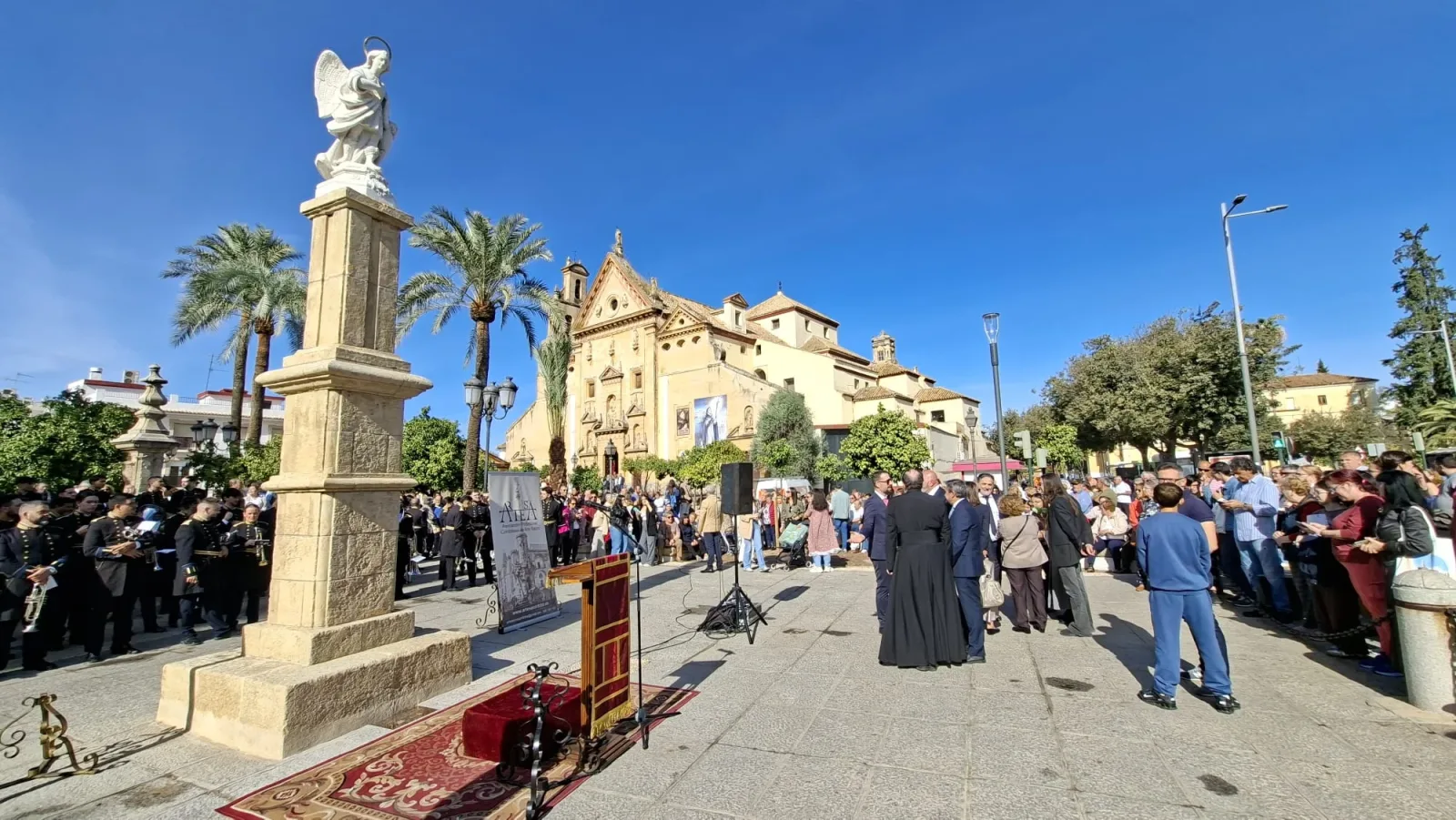 Una multitud congregada en una plaza con una columna rematada por una estatua de un ángel, un atril y una banda de música, ante un edificio religioso de fachada