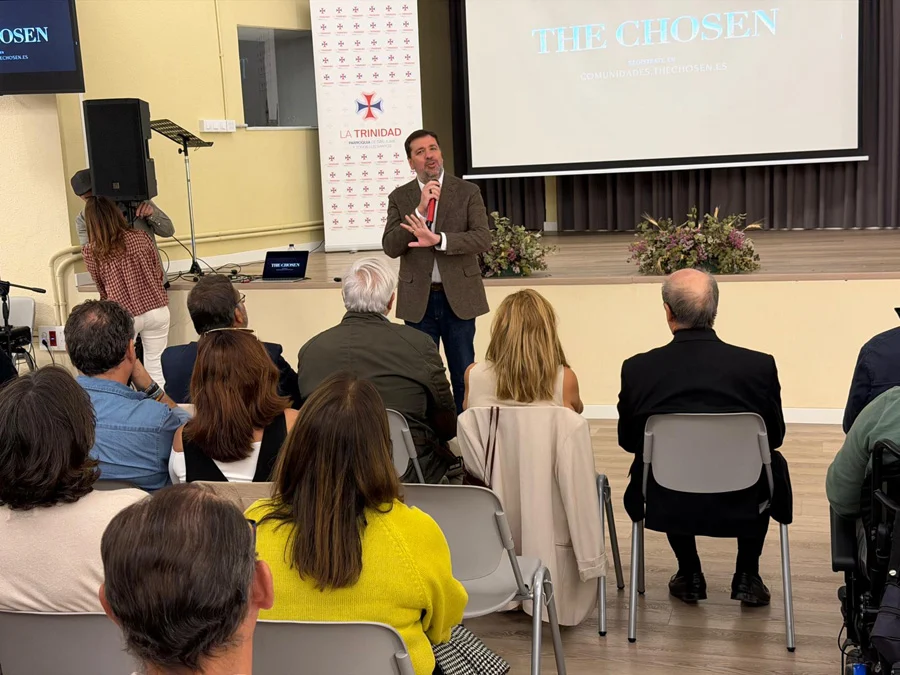 Un hombre con micrófono habla en un escenario ante un público sentado, con una pantalla que muestra The Chosen.