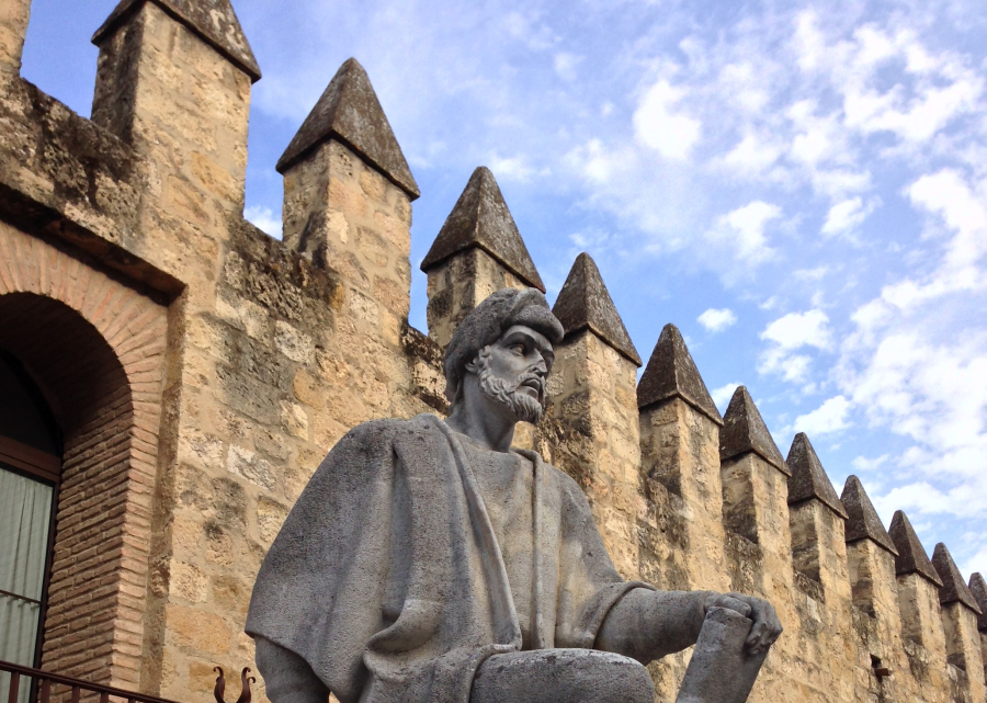 Estatua de piedra de un hombre con turbante y túnica, sentado ante una muralla almenada de piedra bajo un cielo con nubes.