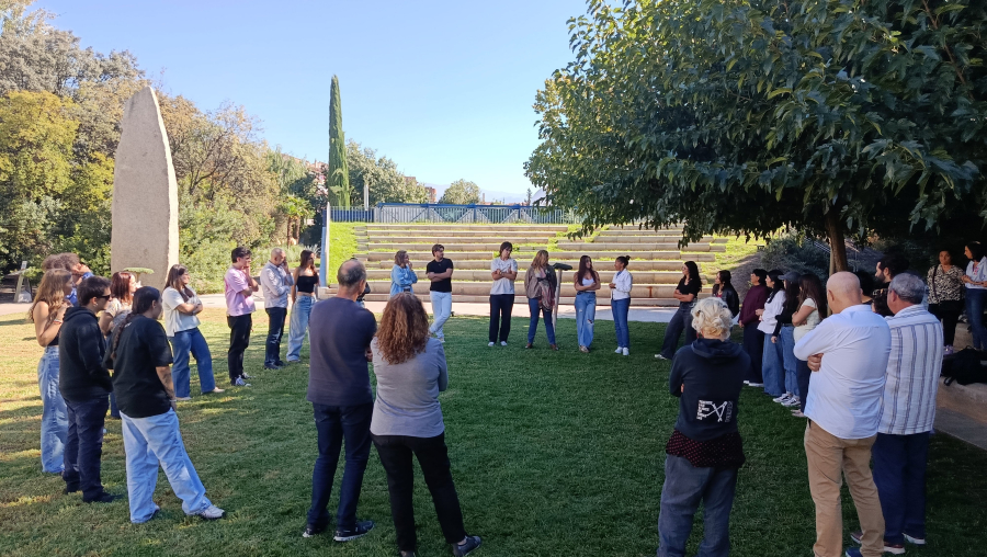 Un grupo de personas de pie en círculo sobre el césped de un parque, junto a unas escaleras y un monumento de piedra.