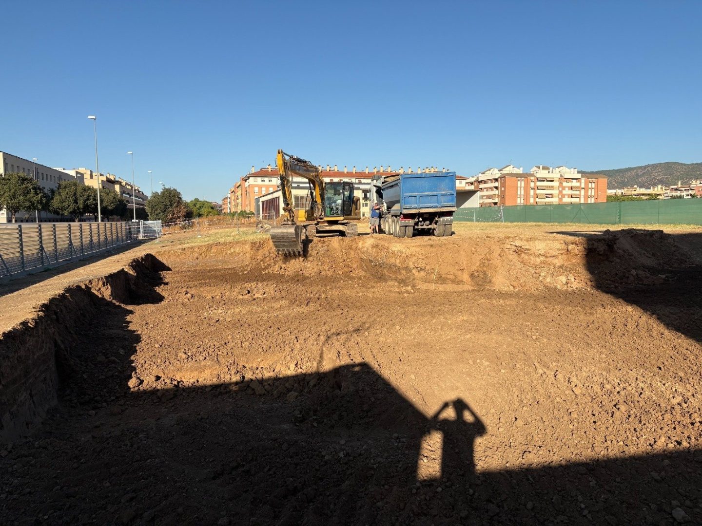Una excavación de tierra con una excavadora amarilla y un camión volquete azul al fondo, junto a edificios y una valla.