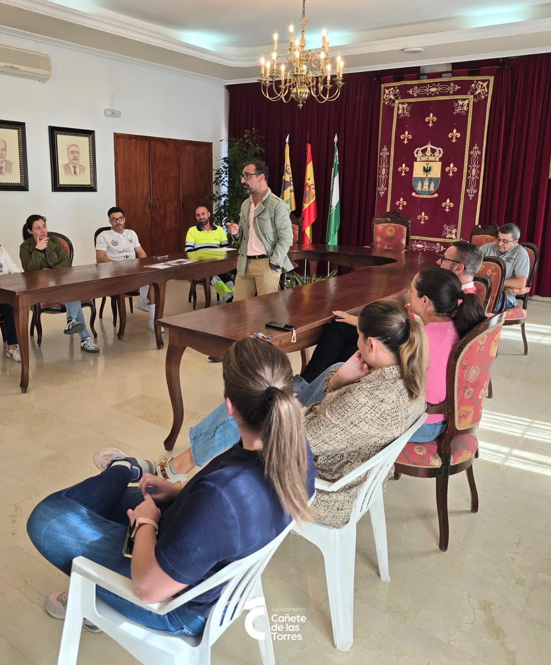 Un hombre de pie habla ante varias personas sentadas alrededor de una mesa de madera en una sala con banderas y un tapiz.