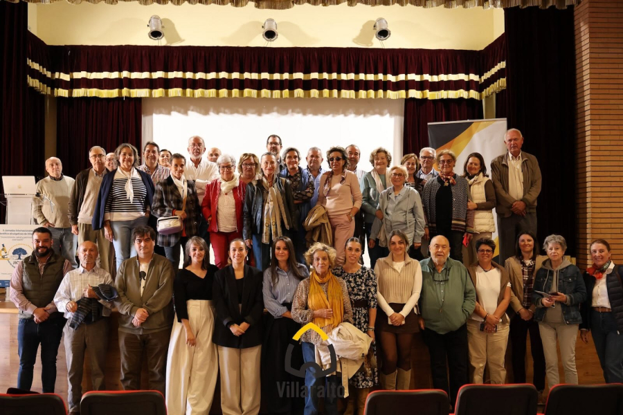 Un grupo numeroso de personas de distintas edades posa de pie en un escenario con cortinas oscuras y un cartel lateral.