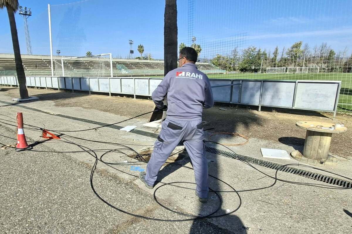 Un operario de espaldas con mono gris trabaja con cables junto a un campo de fútbol, una valla y un cono naranja.