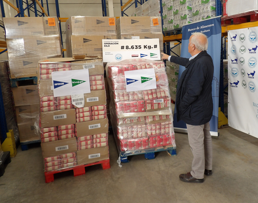 Un hombre mayor señala un cartel sobre dos palés de arroz y galletas envueltos en plástico, en un almacén con estanterías.