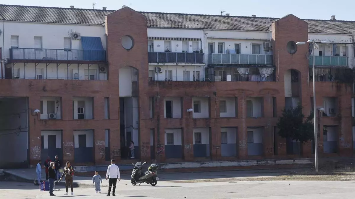 Bloque de viviendas de ladrillo con balcones y locales cerrados, varias personas y una moto aparcada delante.