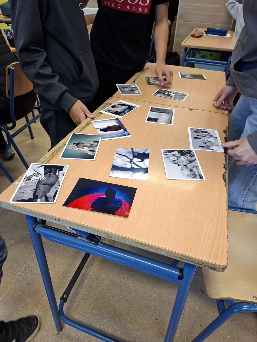 Varias personas de pie junto a una mesa escolar cubierta con fotografías impresas en blanco y negro y en color.