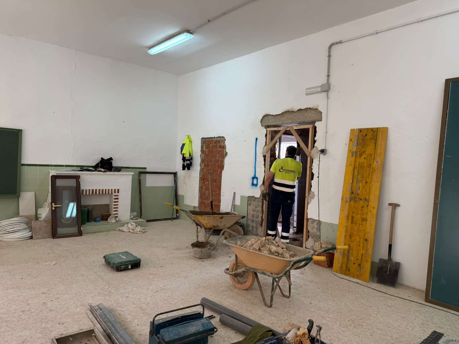 Una habitación amplia en obras con un trabajador junto a un hueco abierto en la pared, dos carretillas con escombros, tablones y herramientas.