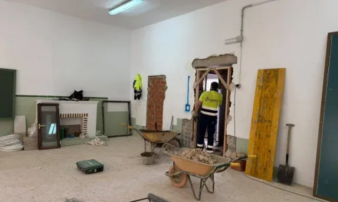 Habitación en obras con un trabajador junto a un hueco abierto en la pared, dos carretillas con escombros y una pizarra verde.