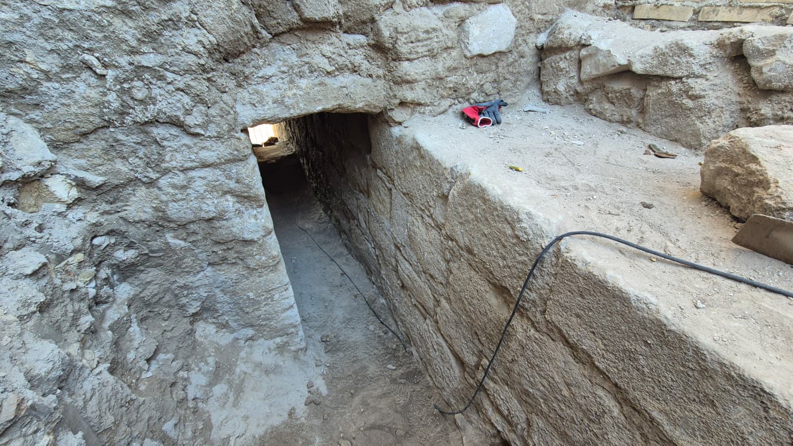 Galería estrecha excavada en roca y muros de piedra, con un cable y guantes sobre una repisa.
