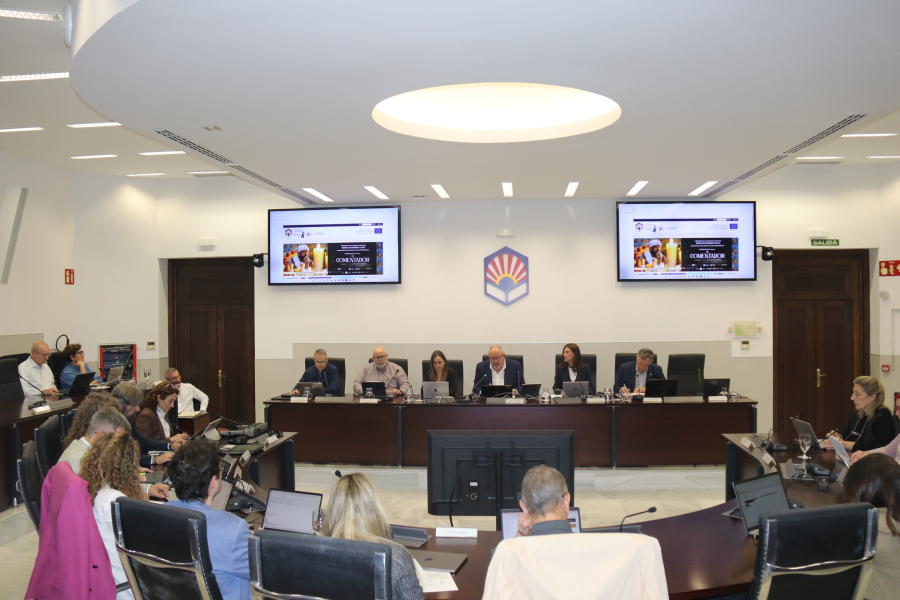 Sala de reuniones con una mesa principal de seis personas y asistentes sentados frente a pantallas y micrófonos.