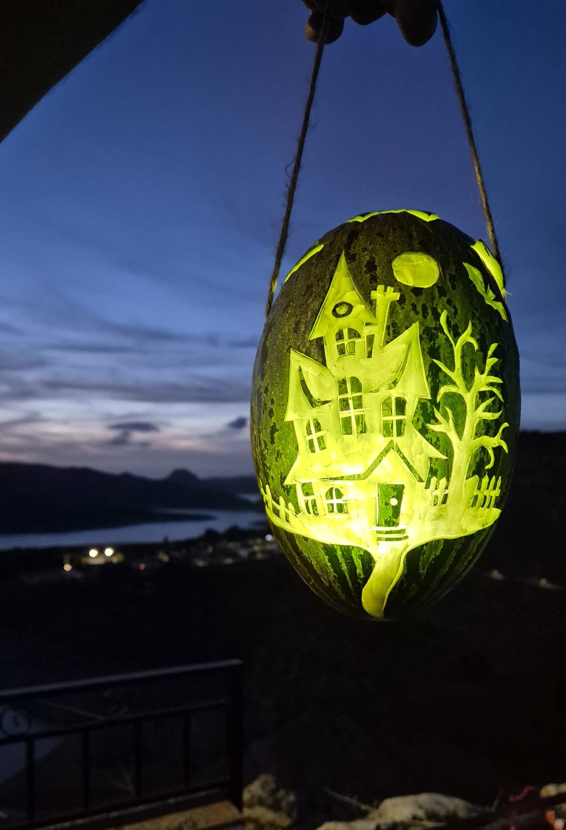 Una sandía colgada y tallada con una casa iluminada por dentro, con un pueblo y un lago al fondo al anochecer.