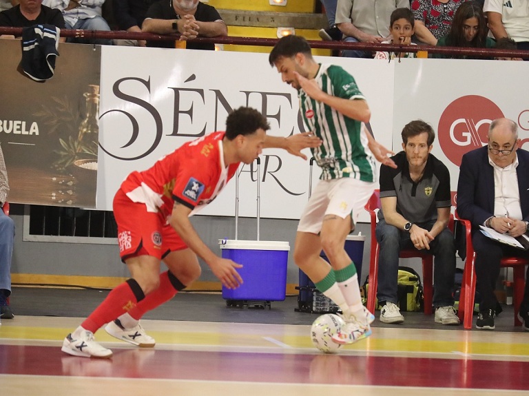 Dos jugadores de fútbol sala, uno con equipación roja y otro con camiseta blanca y verde, disputan el balón junto a la banda.